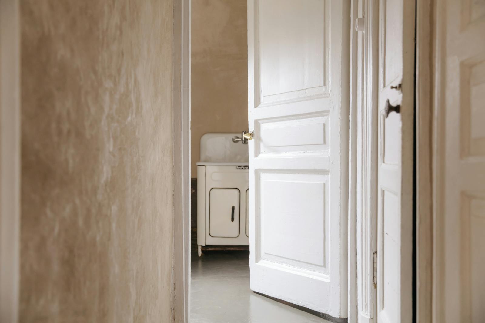 A rustic interior with a white wooden door slightly ajar revealing a vintage sink inside, molds, home